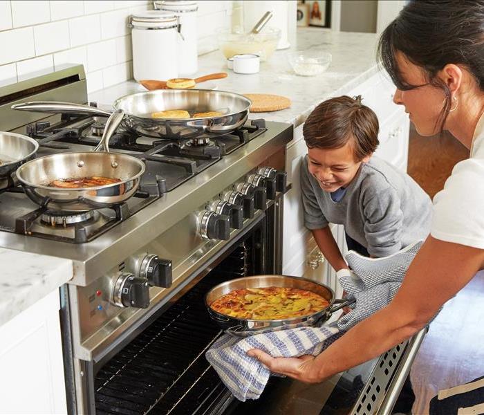 A mother and her son take food out of the oven while other food cooks on the stovetop.